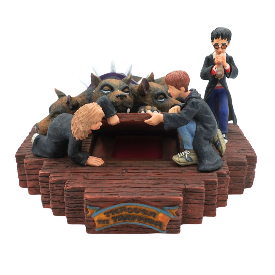 A Harry Potter themed figurine depicting the Three Buckles scene, showing Harry, Ron, and Hermione reaching into the cauldron with three wolves behind them on a wooden platform. A small Harry Potter figure stands on the right.