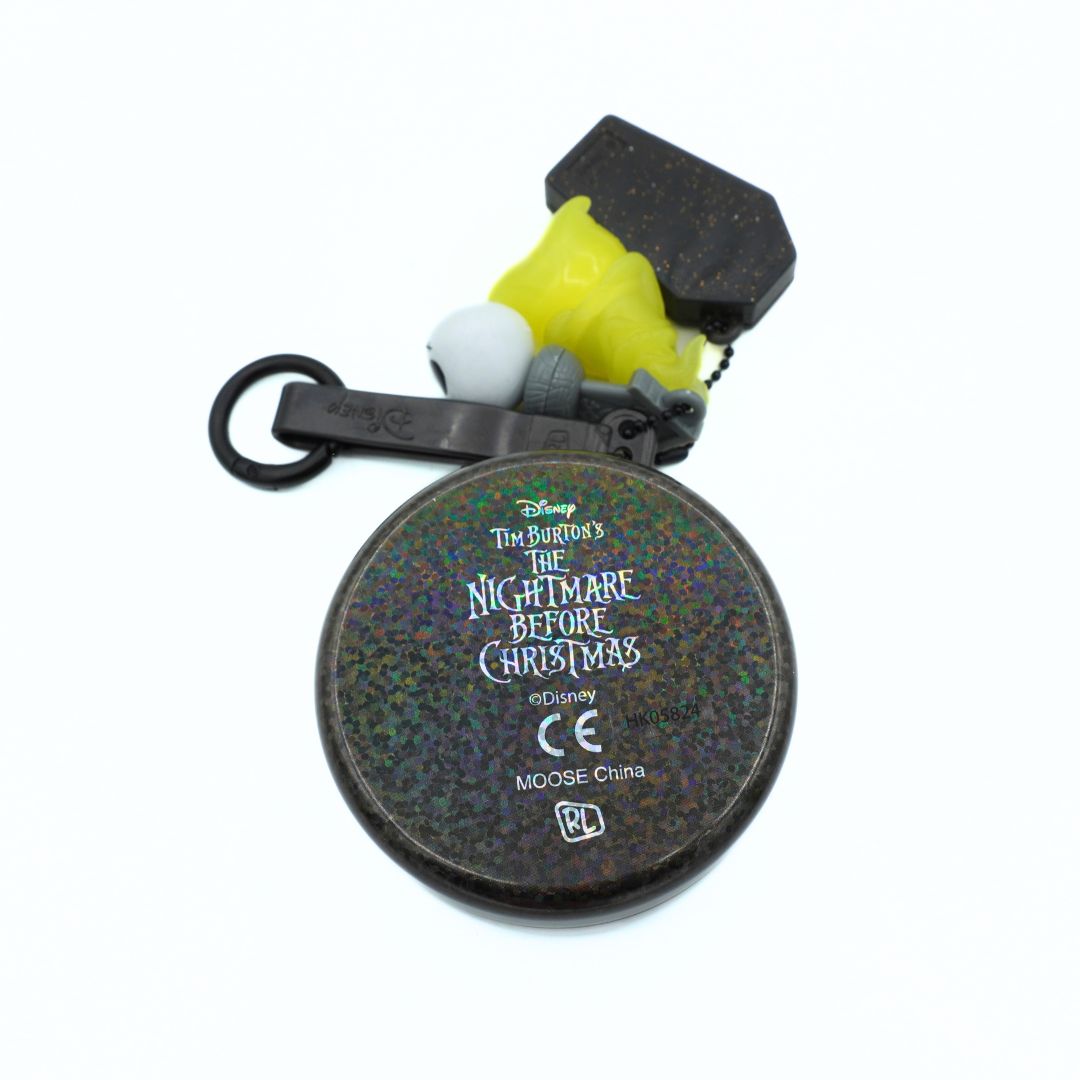 A Disney Tim Burton's The Nightmare Before Christmas themed keychain accessory featuring a round black case with holographic glitter and white text, alongside a black plastic key ring, a white and yellow plastic bird figure, and a black tag.