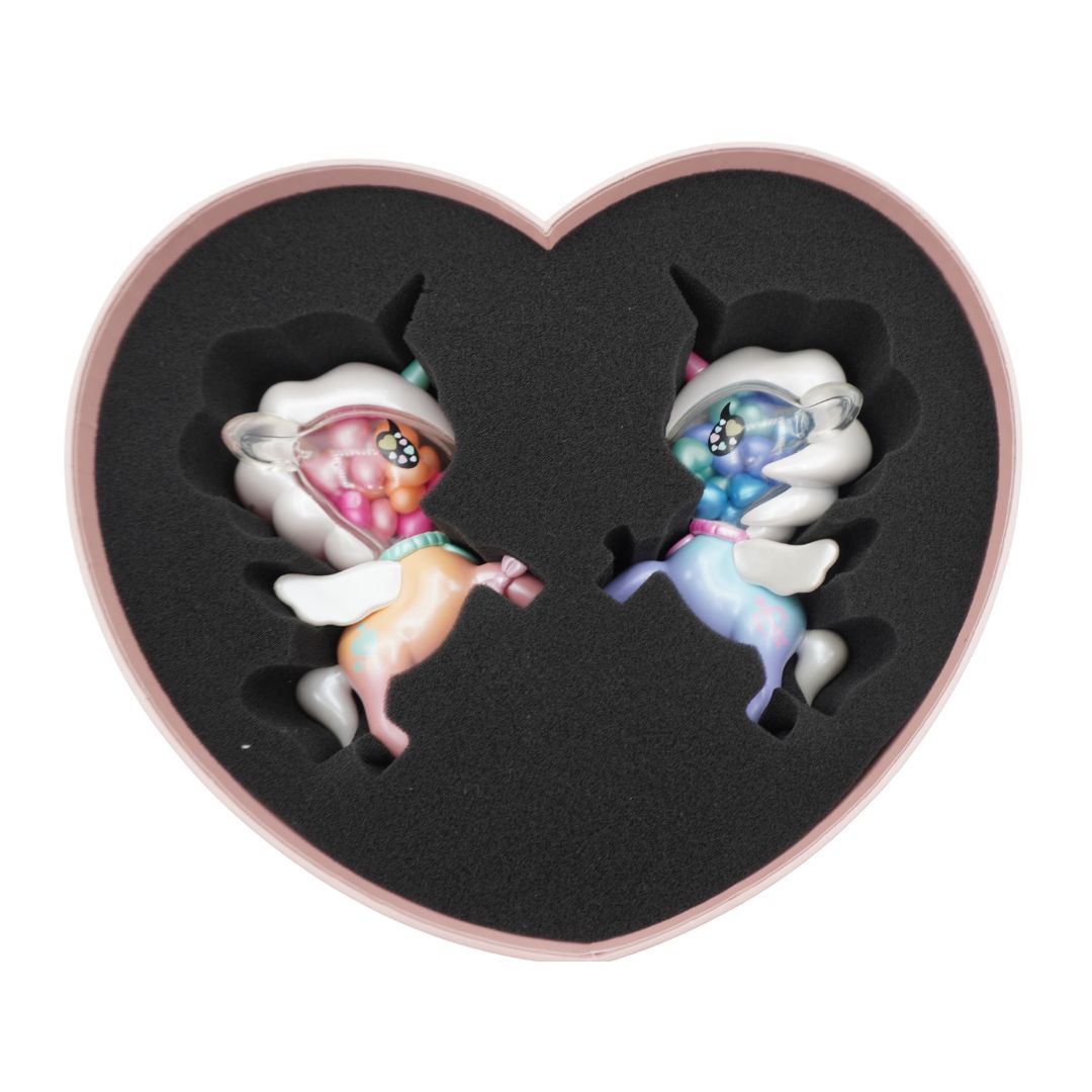 A heart-shaped presentation box with a black foam insert holding two small pastel unicorn figurines facing each other, photographed on a plain white background.