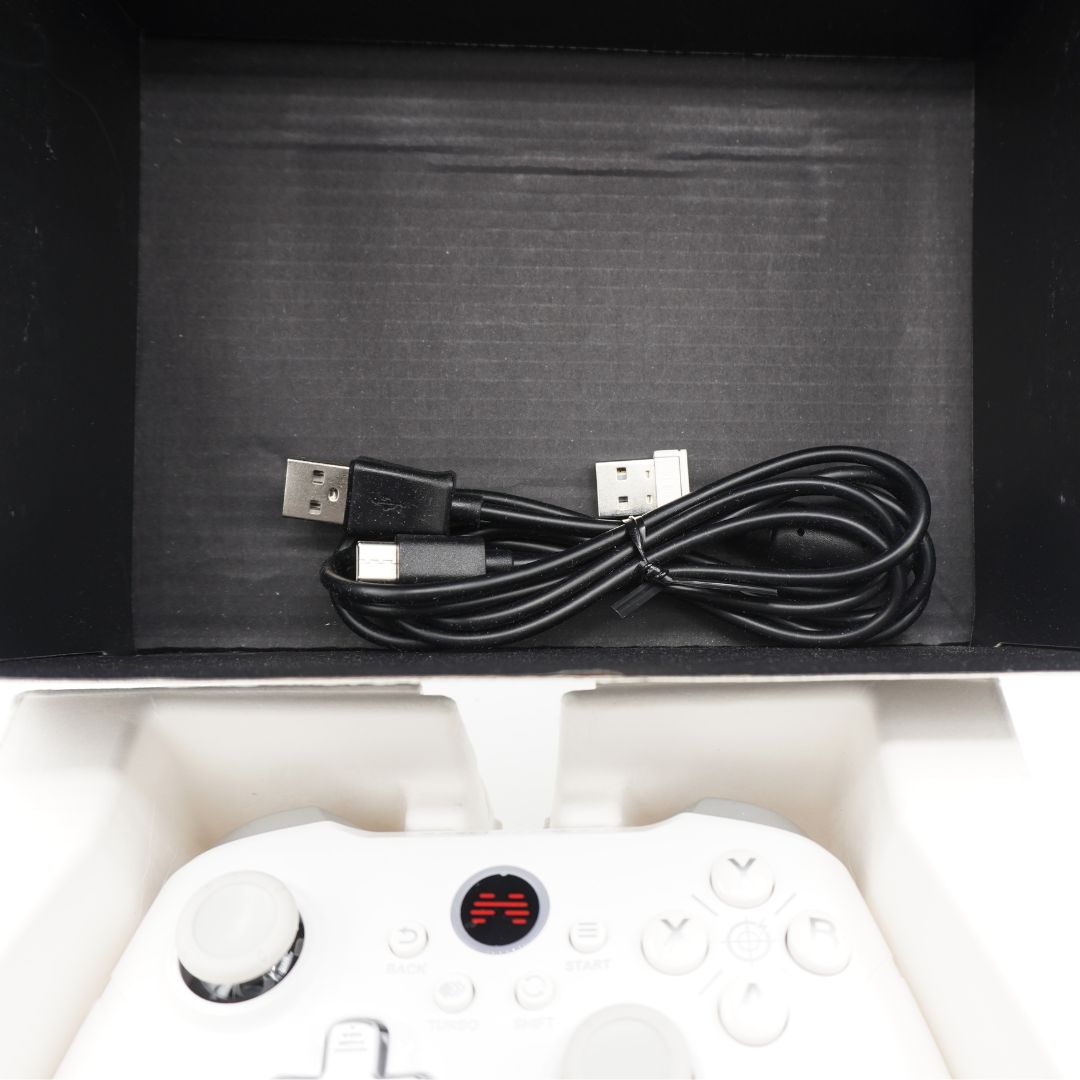 A white game controller is shown in its white foam packaging tray, with a coiled black USB cable resting in the black inner box above it.