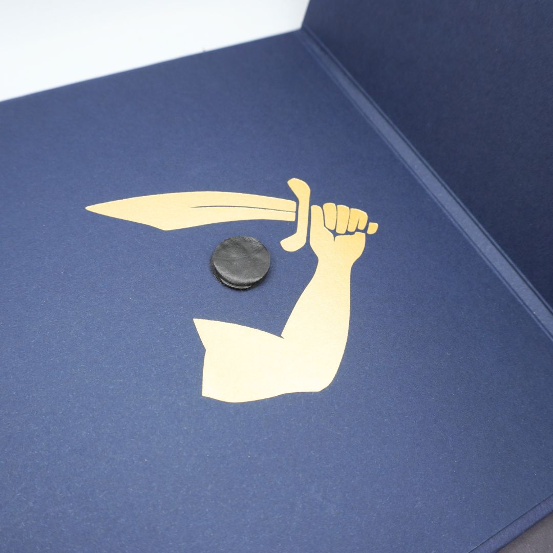 A close-up of a navy blue textured surface featuring a gold foil graphic of a raised arm holding a dagger, with a small round black coin placed in the center.