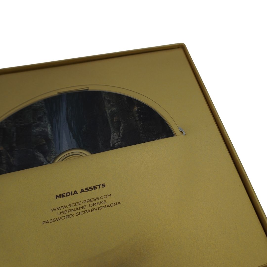 A close-up of a gold-colored media box or DVD case with a partially slid-out cover showing a dark printed insert underneath. Visible text on the cover reads 'MEDIA ASSETS' with website and login/password details.