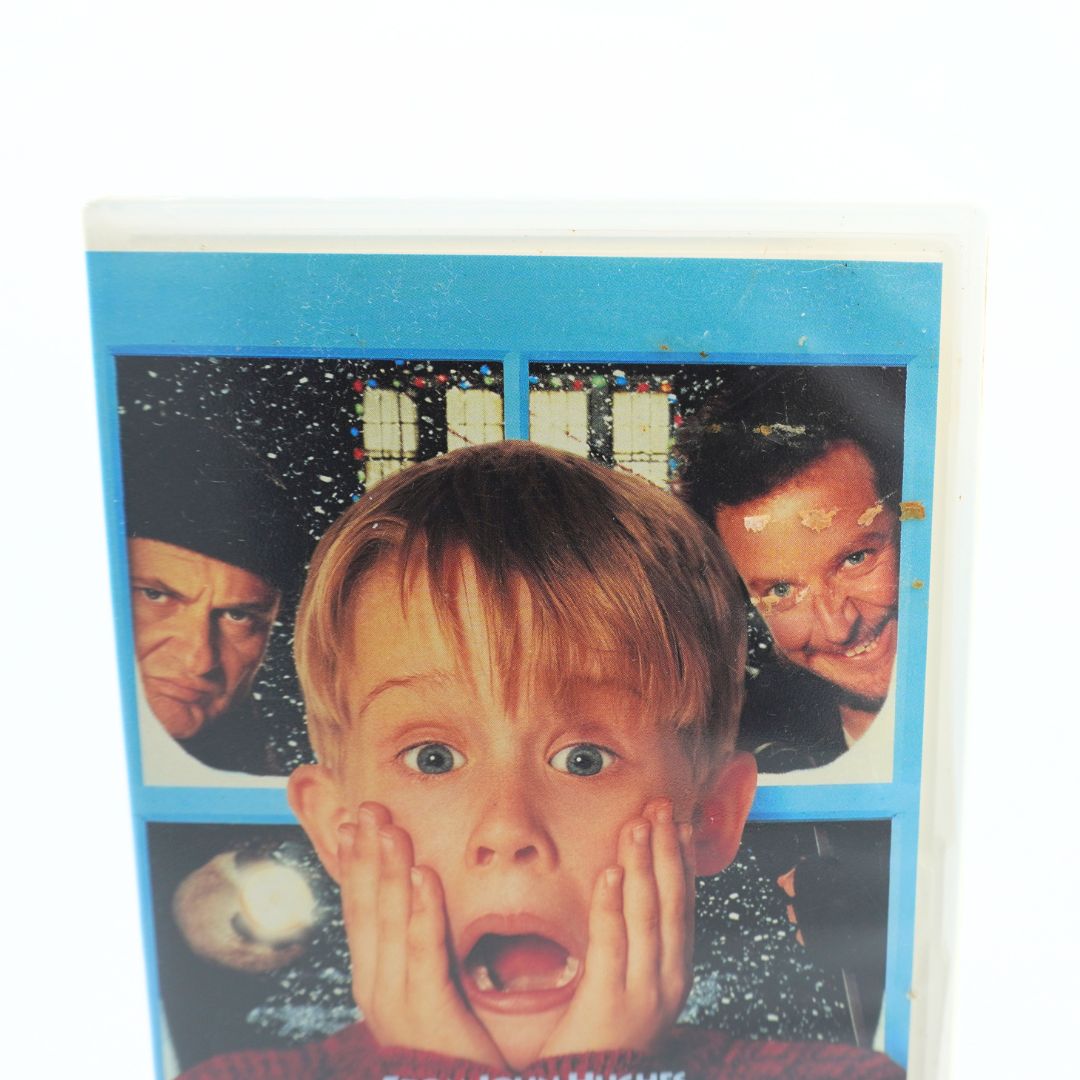 A vintage VHS tape of the movie 'Home Alone' shown against a plain white background. The tape is slightly worn and has a blue border, featuring the main character, Kevin McCallister, in the center with his father and mother pictured in the background panels.