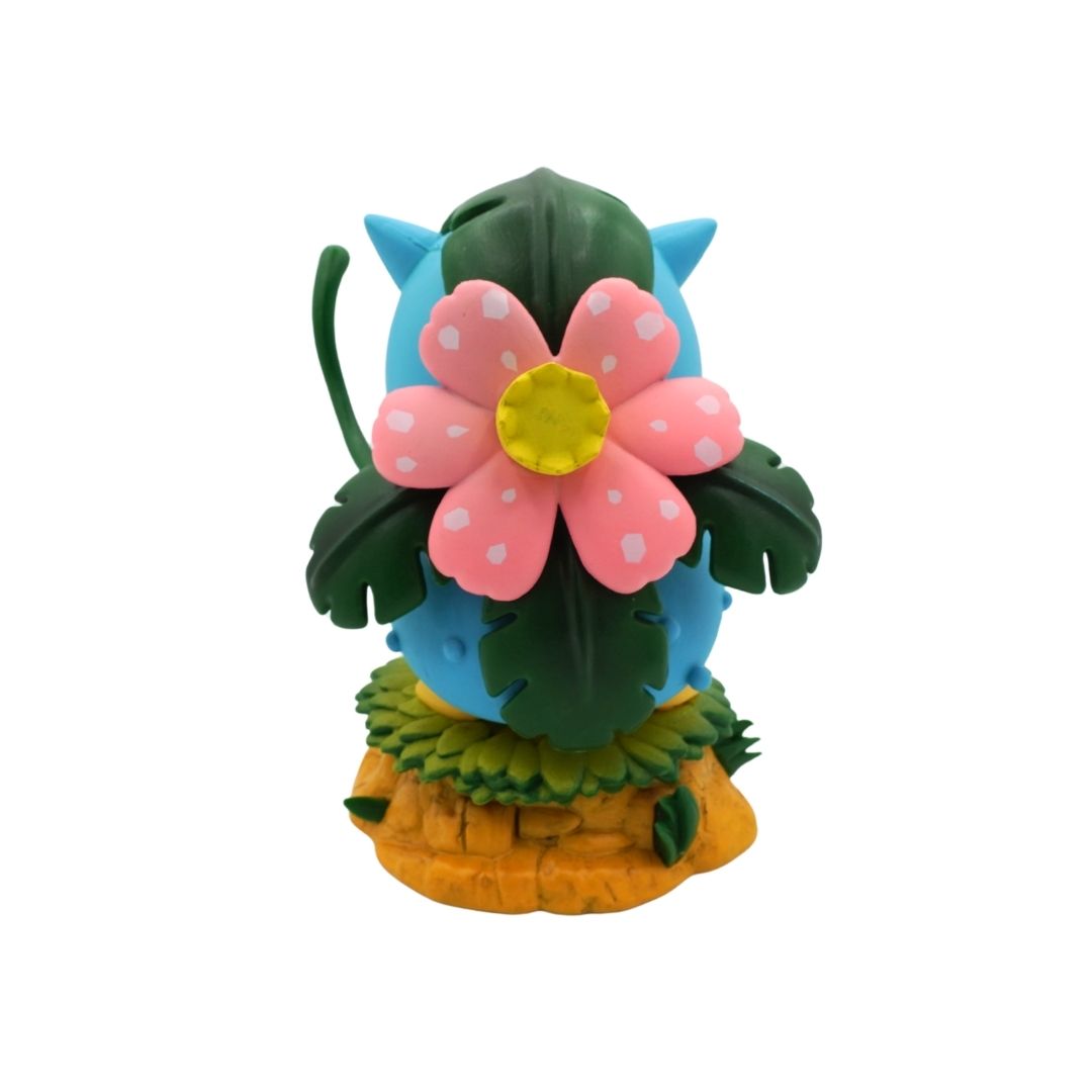 A small collectible figurine of a Venusaur character hidden behind a large pink flower with white spots and a yellow center, set against green leaves on a yellow base, isolated on a white background.