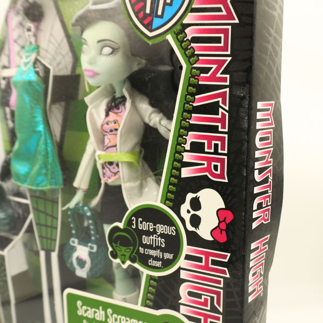 Monster High Sarah Scaramucca doll in its original window-box packaging, featuring a pale green skin tone, black hair, and a white trench coat with a pink and black patterned shirt.