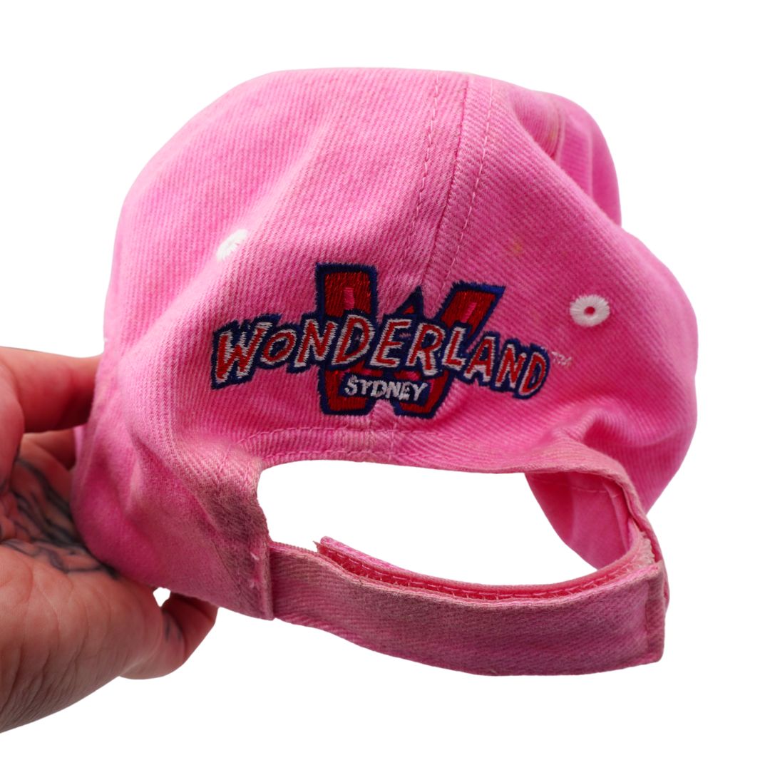 A pink baseball cap held by a hand, featuring an embroidered 'WONDERLAND SYDNEY' logo on the front.