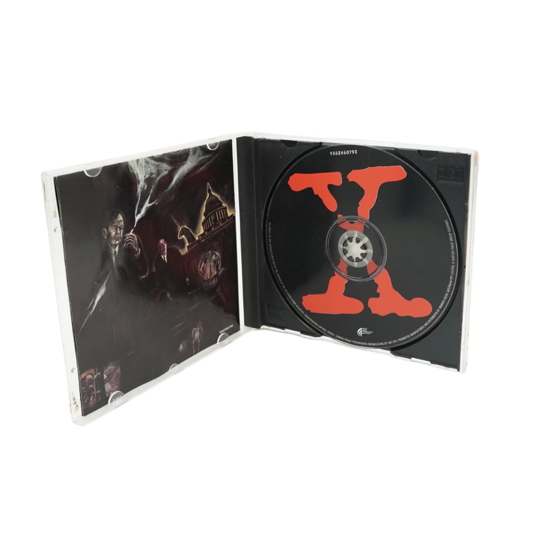 A CD case shown open on a plain white background, with the left inner cover featuring dark illustrated artwork and the right side holding a black CD with a large red stylized letter X.
