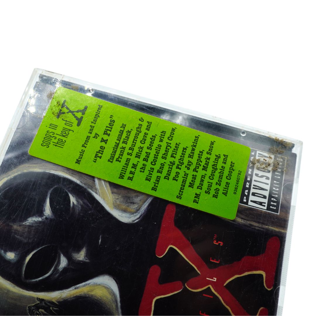 A cassette tape of the 'The X-Files' music album, featuring a bright lime green cover label with tracklist text and a black-and-white illustrated cover with red title lettering.